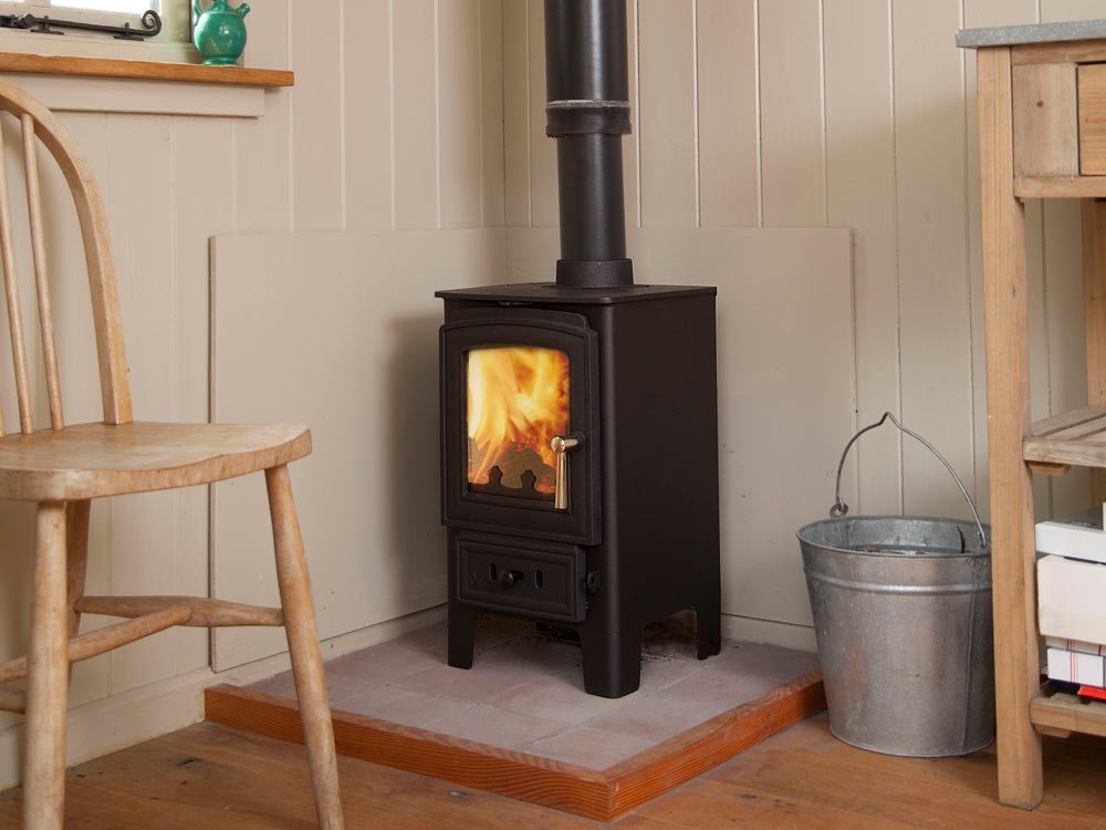 Small black wood burning stove installed in a corner on a stone hearth with metal flue pipe and lit fire